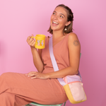 Smiling young woman in a peach sleeveless jumpsuit sitting on a mint green chair against a pastel pink background, holding a yellow ceramic mug with bold text that reads “F*CK THAT” in colorful letters. She wears a pastel color-blocked crossbody bag in peach, lilac, and yellow tones with a wide lavender strap, and has a sun tattoo on her upper arm and statement star earrings.
