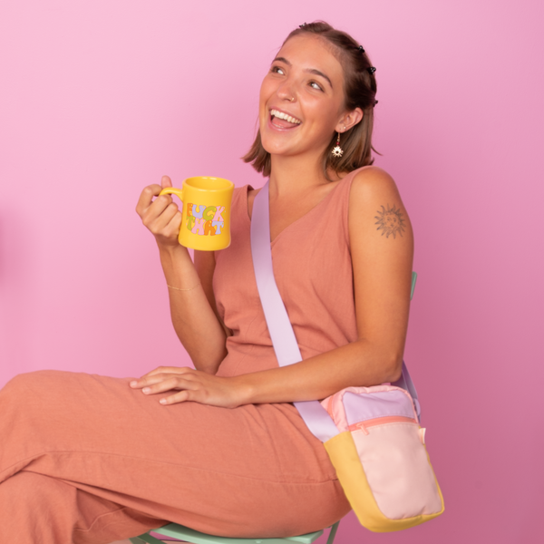 Smiling young woman in a peach sleeveless jumpsuit sitting on a mint green chair against a pastel pink background, holding a yellow ceramic mug with bold text that reads “F*CK THAT” in colorful letters. She wears a pastel color-blocked crossbody bag in peach, lilac, and yellow tones with a wide lavender strap, and has a sun tattoo on her upper arm and statement star earrings.