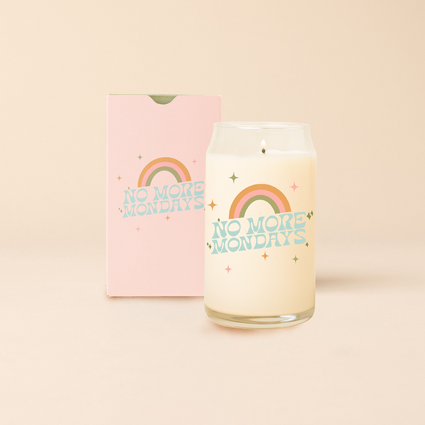 Can glass candle with text that reads "No More Mondays" in multi color font, with minimalist sparkle stars surrounding text, 3 stripe rainbow sits above text. Box packaging with the same design sits behind candle.