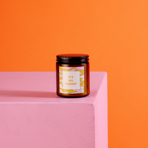 Amber glass candle jar with sticker on front with multi-color wavy and floral design and text that reads "IT'S ALL GROOVY" in orange font in a white square.