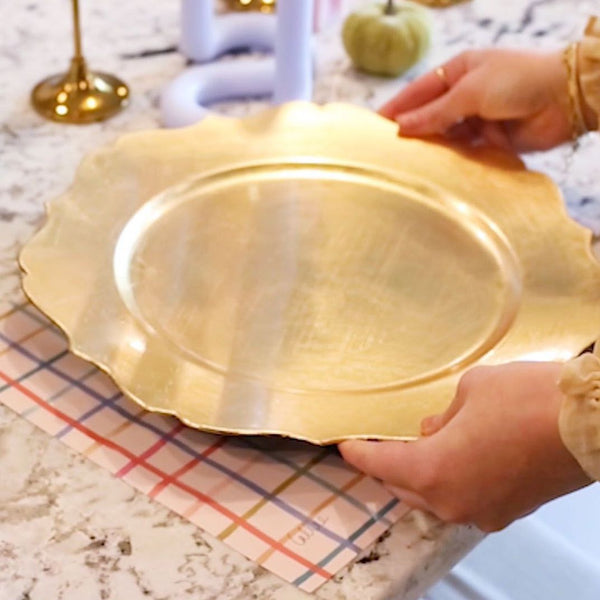 Pastel Plaid Placemat in Table Setting
A pastel plaid placemat featuring intersecting lines in vibrant hues of pink, orange, blue, and olive green sits on a marble countertop. A gold scalloped charger plate is being gently placed on top of the placemat, partially covering the pattern. The setting evokes a modern, cheerful, and festive table décor.