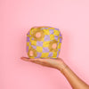A picture of a person's hand holding a cute puffy pouch with a warped checkerboard pattern in citron and periwinkle with camel colored daisies scattered about on a blush pink background.