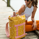 Model with a mustard yellow colored cooler backpack. Designed with abstract women faces in white coloring. Pink detailing on the front and straps of the bag.