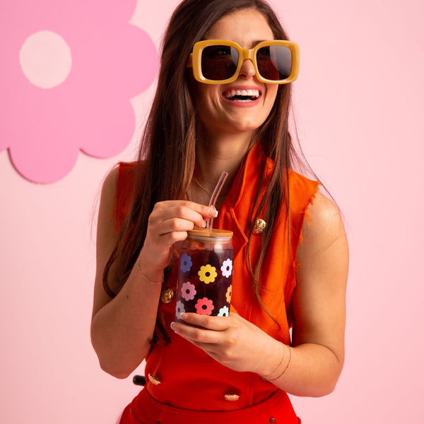 "A girl wearing an orange dress 
with fun retro sunnies holding a clear can glass tumbler filled with iced coffee. The can glass tumbler has a lid and a clear glass straw and has multi colored daisy's scattered all around.