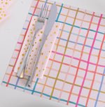 Colorful Table Setting with Checkered Placemat
A modern table setting featuring a pink placemat with a multicolor grid pattern in red, orange, gold, teal, and blue. A shiny fork and knife rest on pink napkins decorated with small gold geometric shapes, adding a festive and cheerful touch.