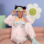 Smiling girl in pastel pink pjs and a checkered sleep mask that says Fuck Off, also wearing a checkered Neck Wrap. 