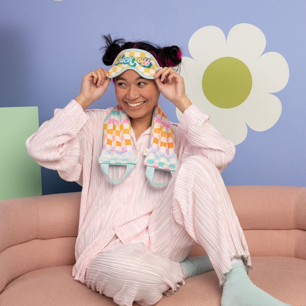 Smiling girl in pastel pink pjs and a checkered sleep mask that says Fuck Off, also wearing a checkered Neck Wrap. 